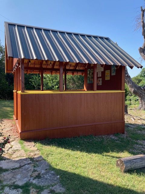Small wooden kiosk with a striped metal roof, standing outdoors in a grassy area.