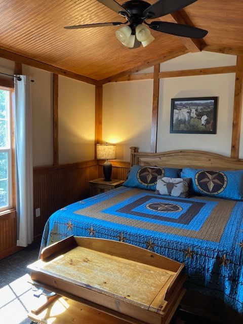 Cozy bedroom with blue quilted bed, wooden tray table, lamp, ceiling fan, and framed wall art.