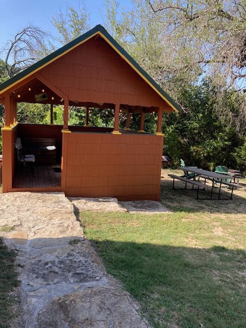 Wooden pavilion with a red roof beside a stone path and picnic tables in a grassy park