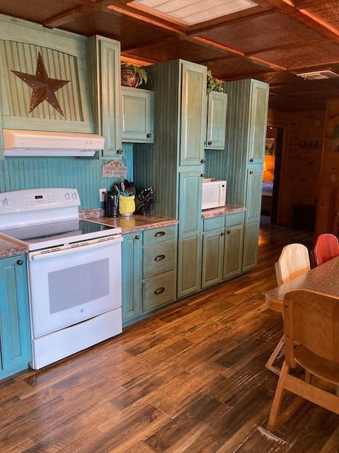 Rustic kitchen with teal cabinets, white appliances, wood floors, and a wooden dining chair
