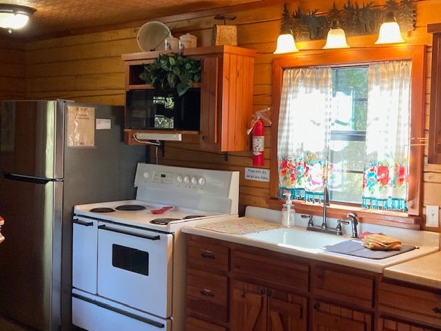 Cozy log cabin kitchen with white stove, stainless refrigerator, wooden cabinets, and sunlit window above sink