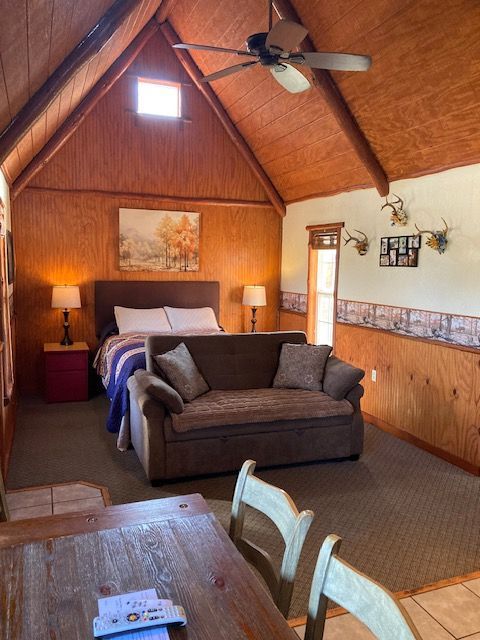 Cozy wood-paneled cabin bedroom with a bed, sofa, ceiling fan, and dining table in foreground