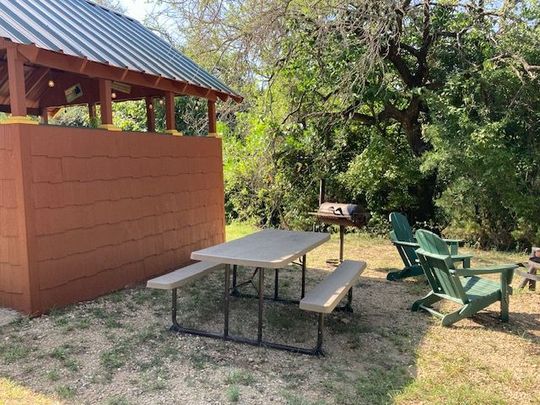 Picnic table and benches beside a brick building, with green chairs in a sunny yard.