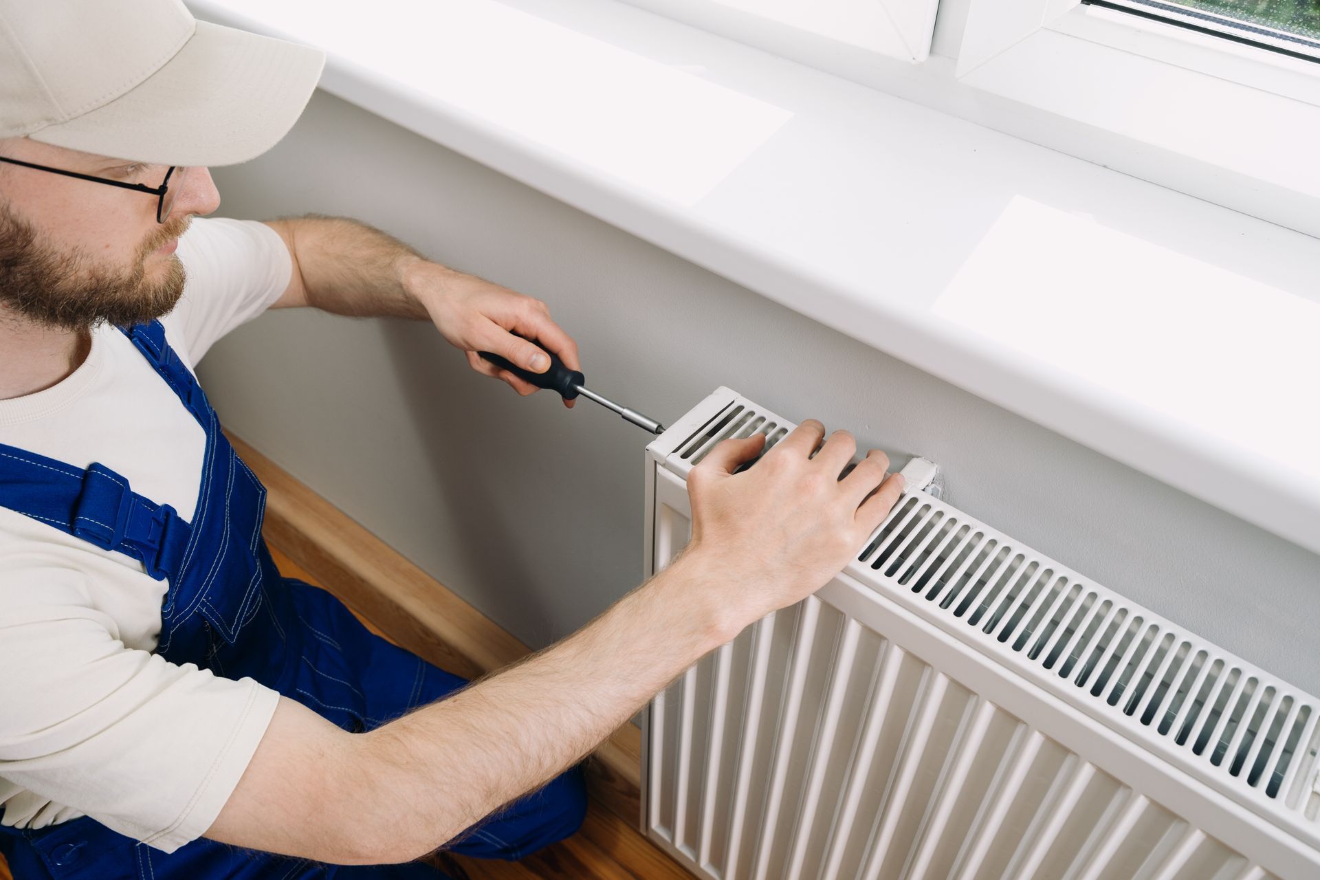 Technician performing heating system repair on white radiator near window with screwdriver. Technician performing heating system repair on white radiator near window with screwdriver.