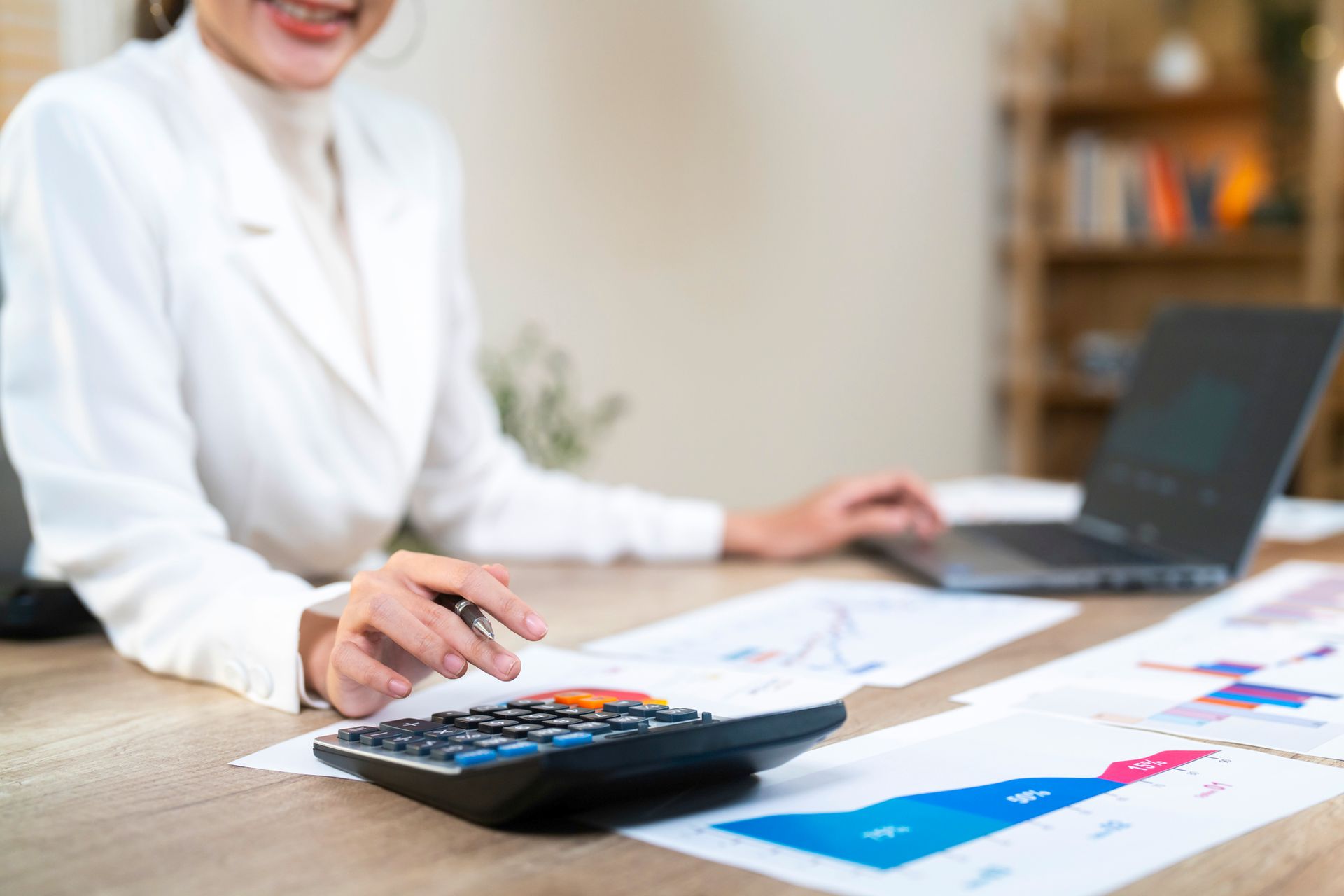 Woman using calculator with charts and laptop on desk.