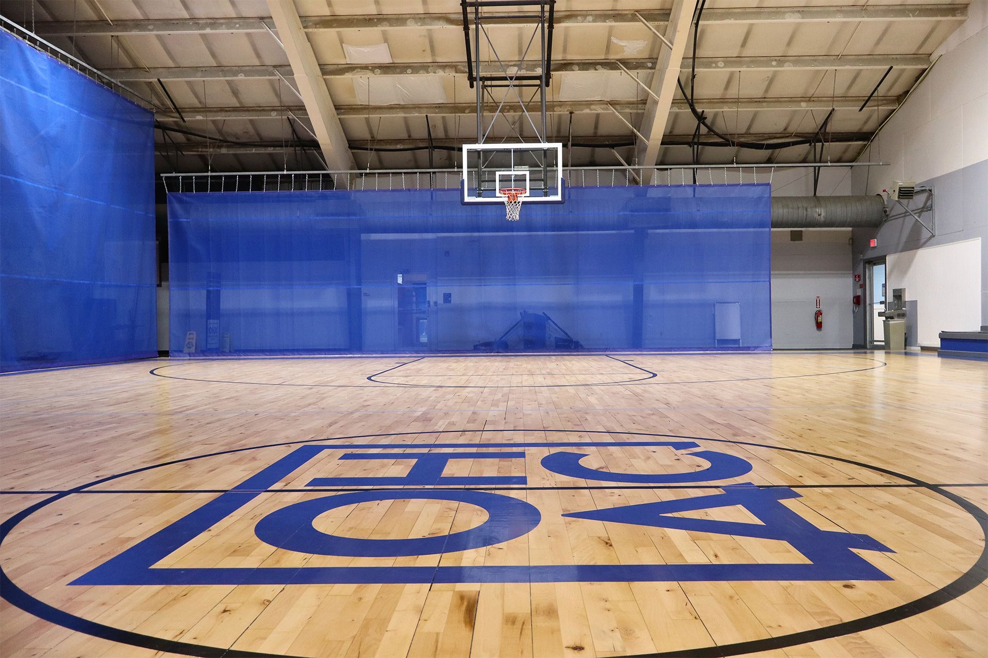 Indoor basketball court with hoop, blue wall padding, and large blue logo on the floor.