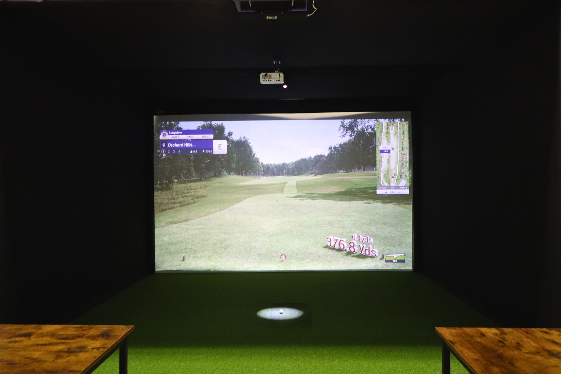 Dim golf simulator room with a large screen showing a golf fairway.