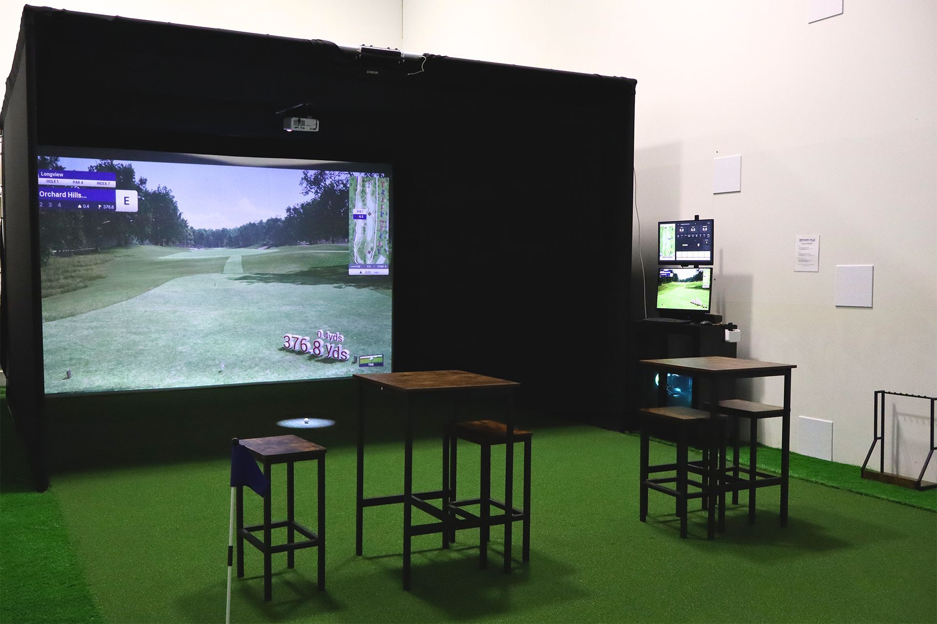 Indoor gaming setup with a large projected racing game, stools, tables, and a monitor on a green-carpeted floor.