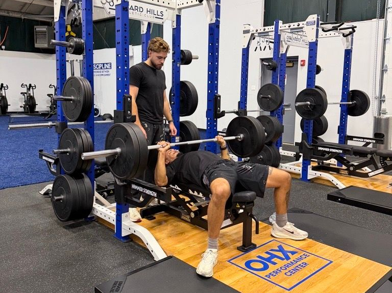 Man bench pressing in a gym, with a spotter standing behind and weight plates on the bar.