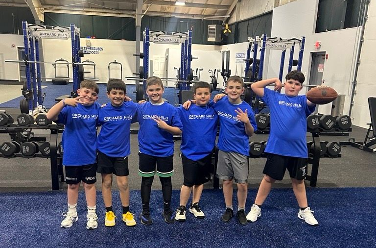 Six boys in blue shirts pose in a gym, standing in front of weightlifting racks and equipment.