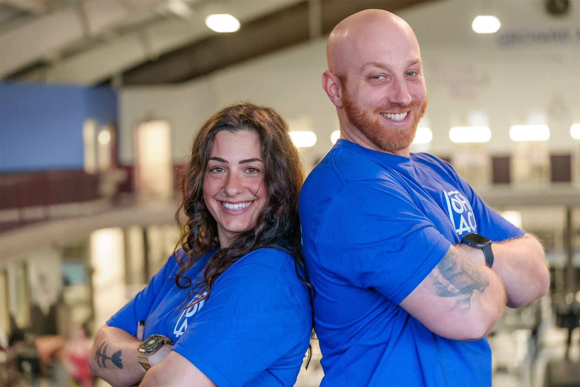 Two smiling coworkers in blue shirts standing back-to-back in a gym or warehouse setting