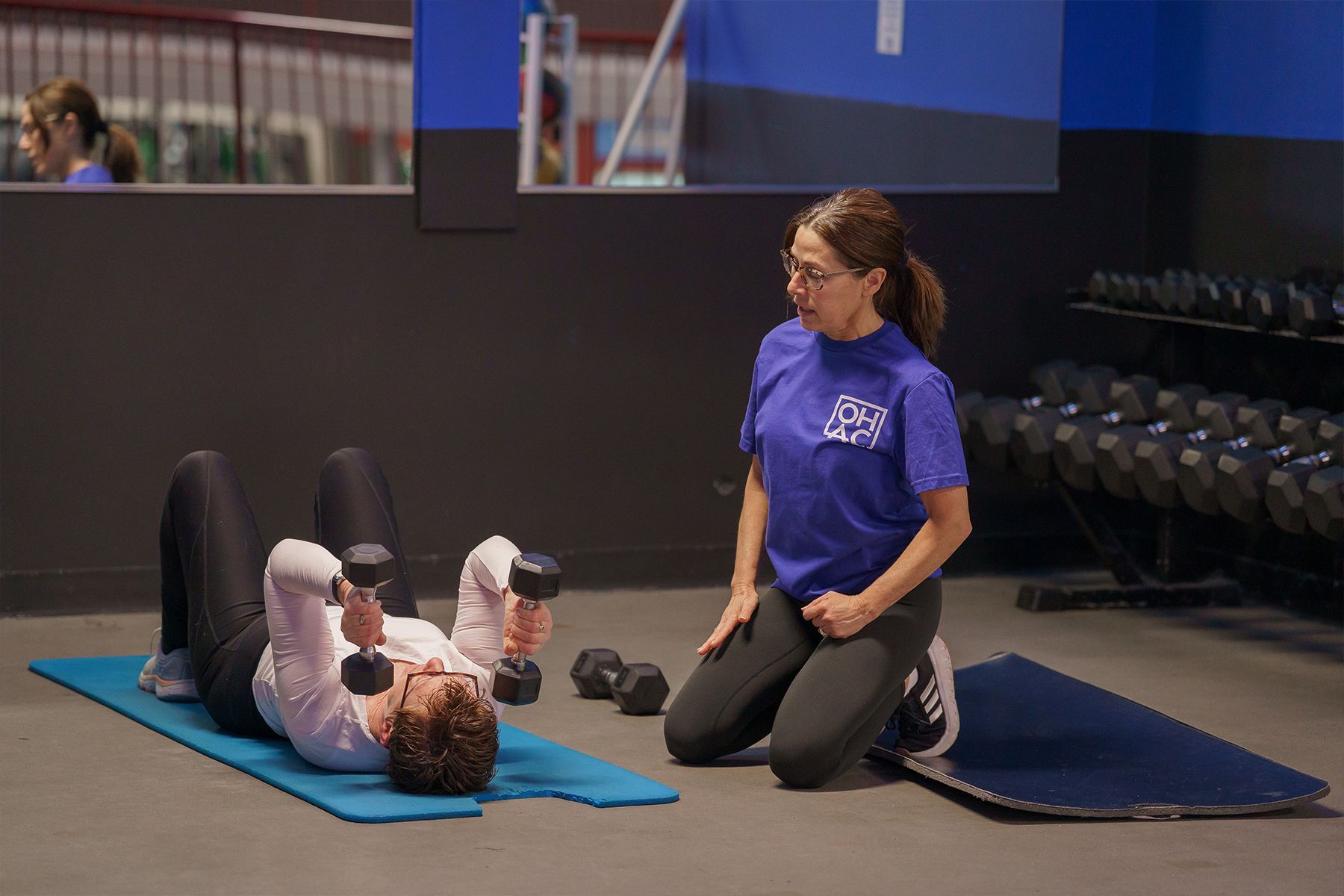 A woman is kneeling down next to a woman lifting dumbbells in a gym.