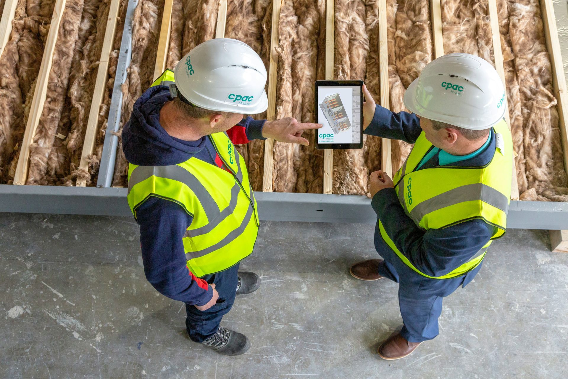 Two workers in hard hats and safety vests reviewing a tablet at a construction site.