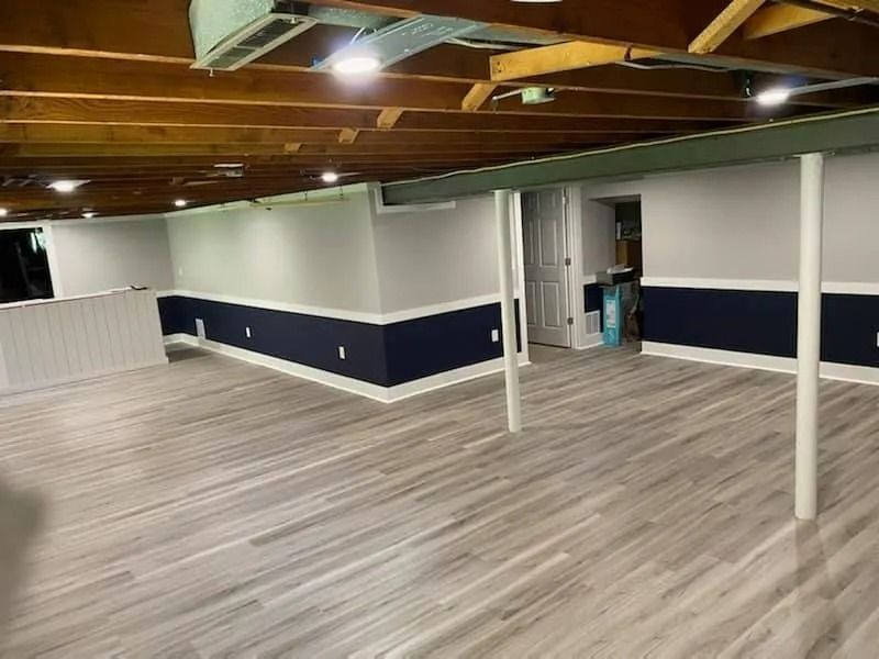 Empty basement room with wood floor, exposed ceiling beams, and navy-and-white walls