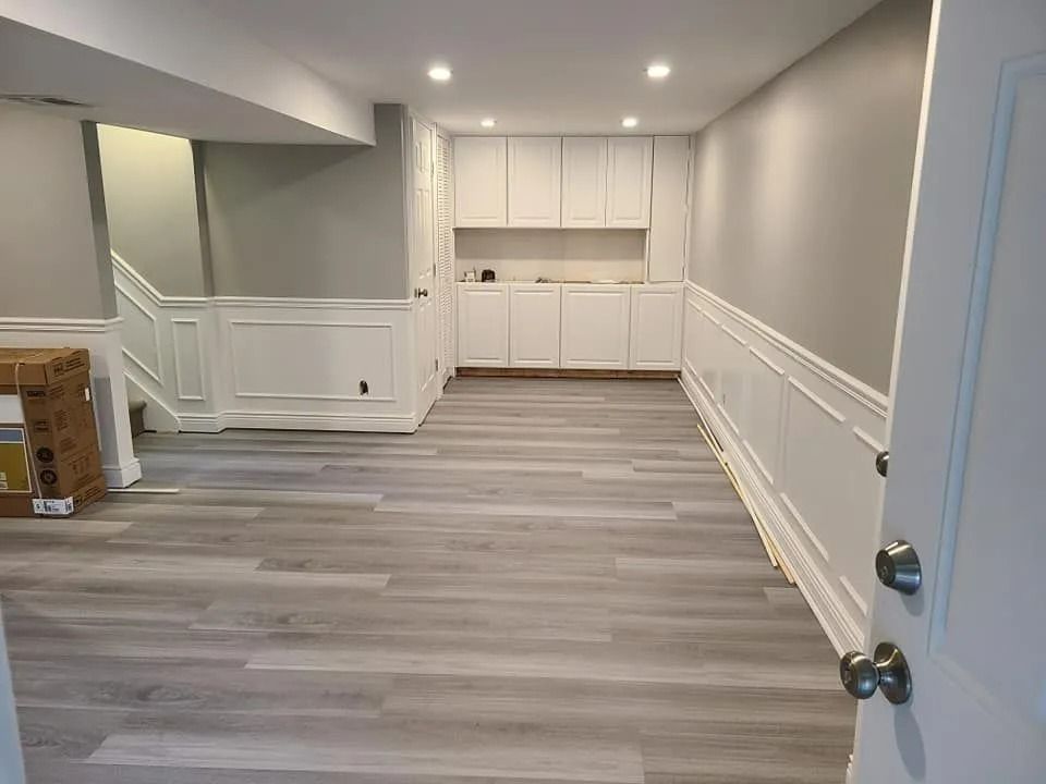 Empty finished basement with gray wood-look floors, white wainscoting, and built-in cabinets at the back.
