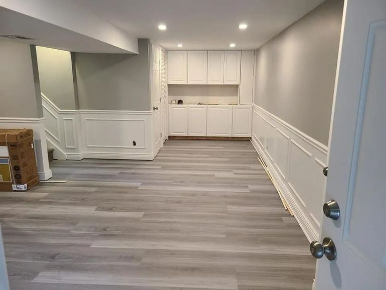 Empty modern room with gray wood floors, white wainscoting, recessed lights, and built-in cabinets at the back