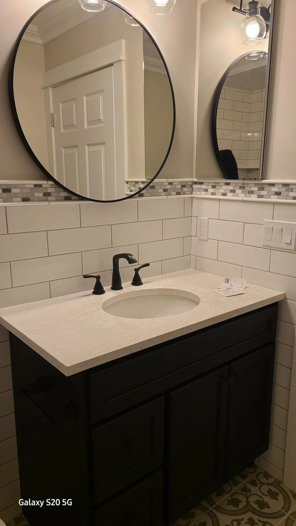 Bathroom vanity with white sink, black cabinet, circular mirrors, and tiled backsplash with patterned floor