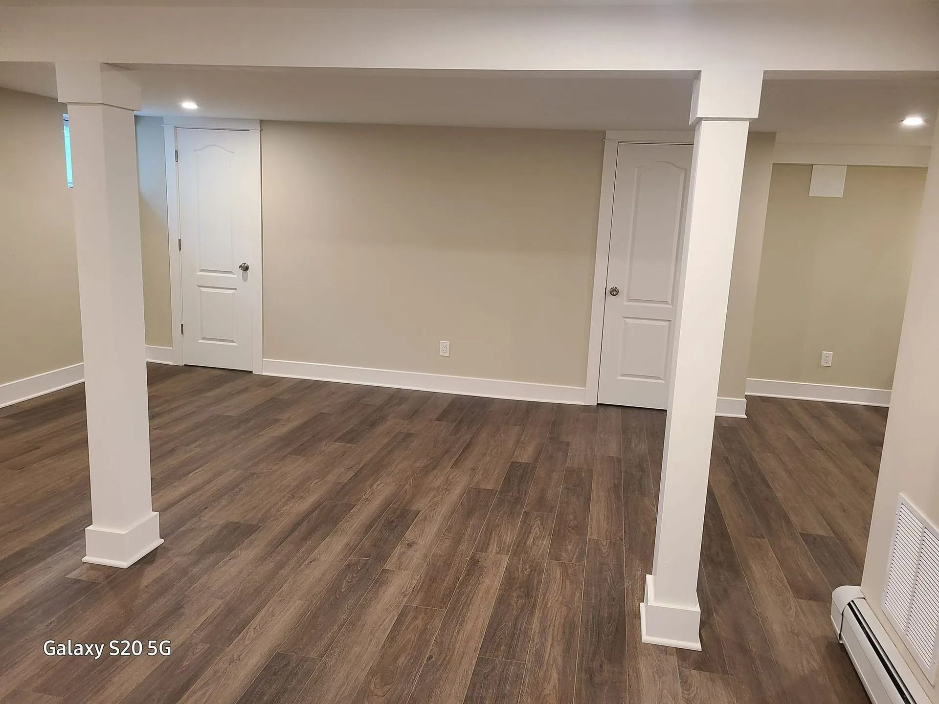 Empty beige room with wood flooring, white columns, and doors on the back wall.