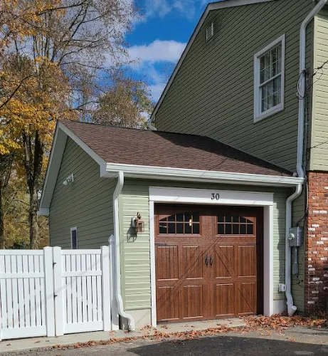 Small light-green garage with a brown door beside a white fence and house under a blue sky.