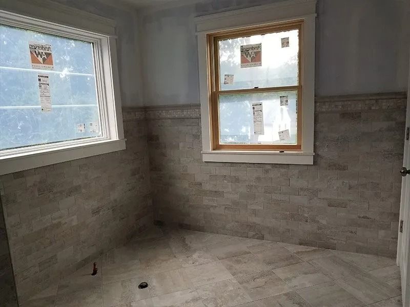 Empty unfinished bathroom with gray tile walls, bare floor, and two windows.