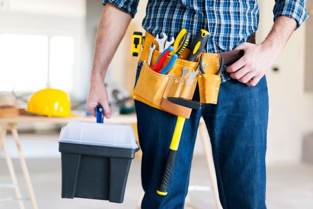 Person holding a toolbox and wearing a tool belt with tools in a bright indoor workspace
