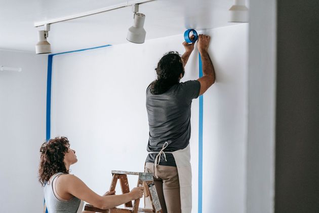 Two people painting a white wall, one on a ladder and one taping the edges inside a room.
