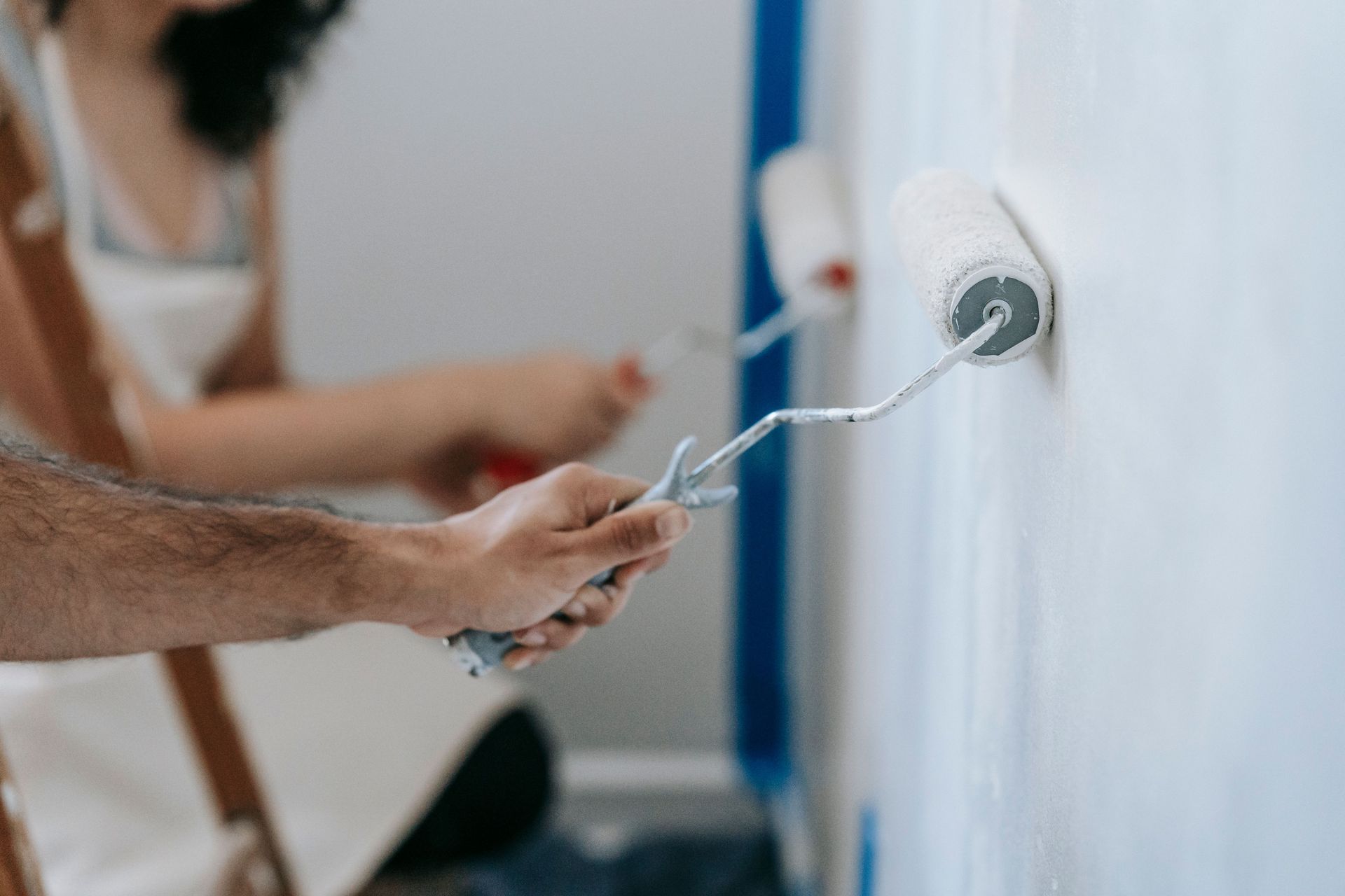 Hands painting a white wall with a paint roller while another person works in the background