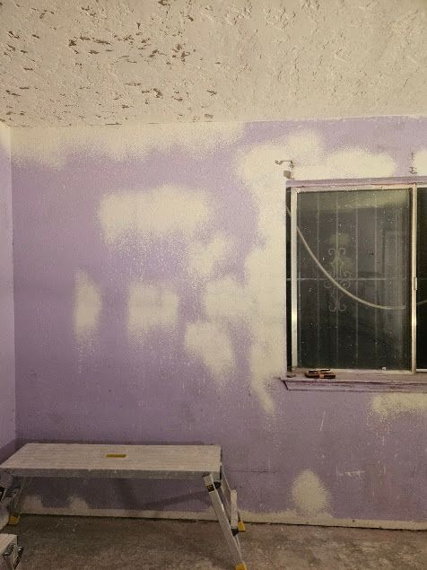 Purple-painted wall with patchy paint, a window, and a small bench in a sparse room.