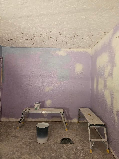 Small room with purple walls, unfinished ceiling, workbench, step stool, paint bucket, and patchy wall repairs.