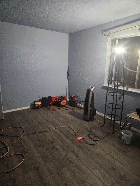 Empty room with gray walls, wood floor, tools, cords, and a bright work light near the window.
