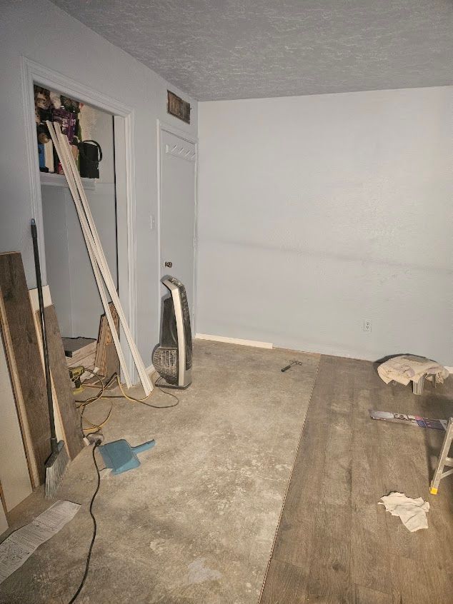 Empty unfinished room with exposed flooring, white walls, and construction materials along the left side.