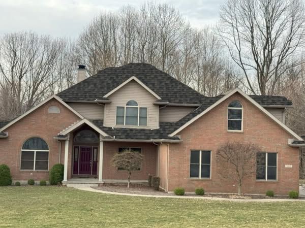 A large brick house with a black roof is surrounded by trees.