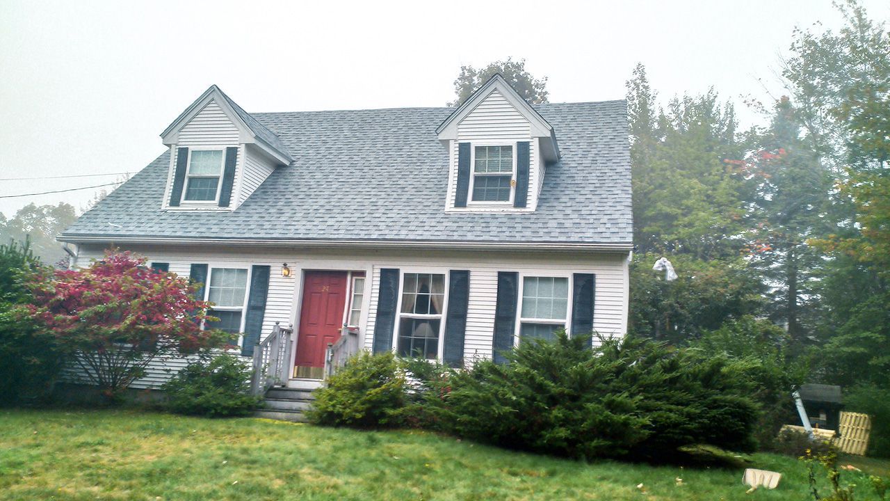 A white house with a blue roof and black shutters is surrounded by trees.