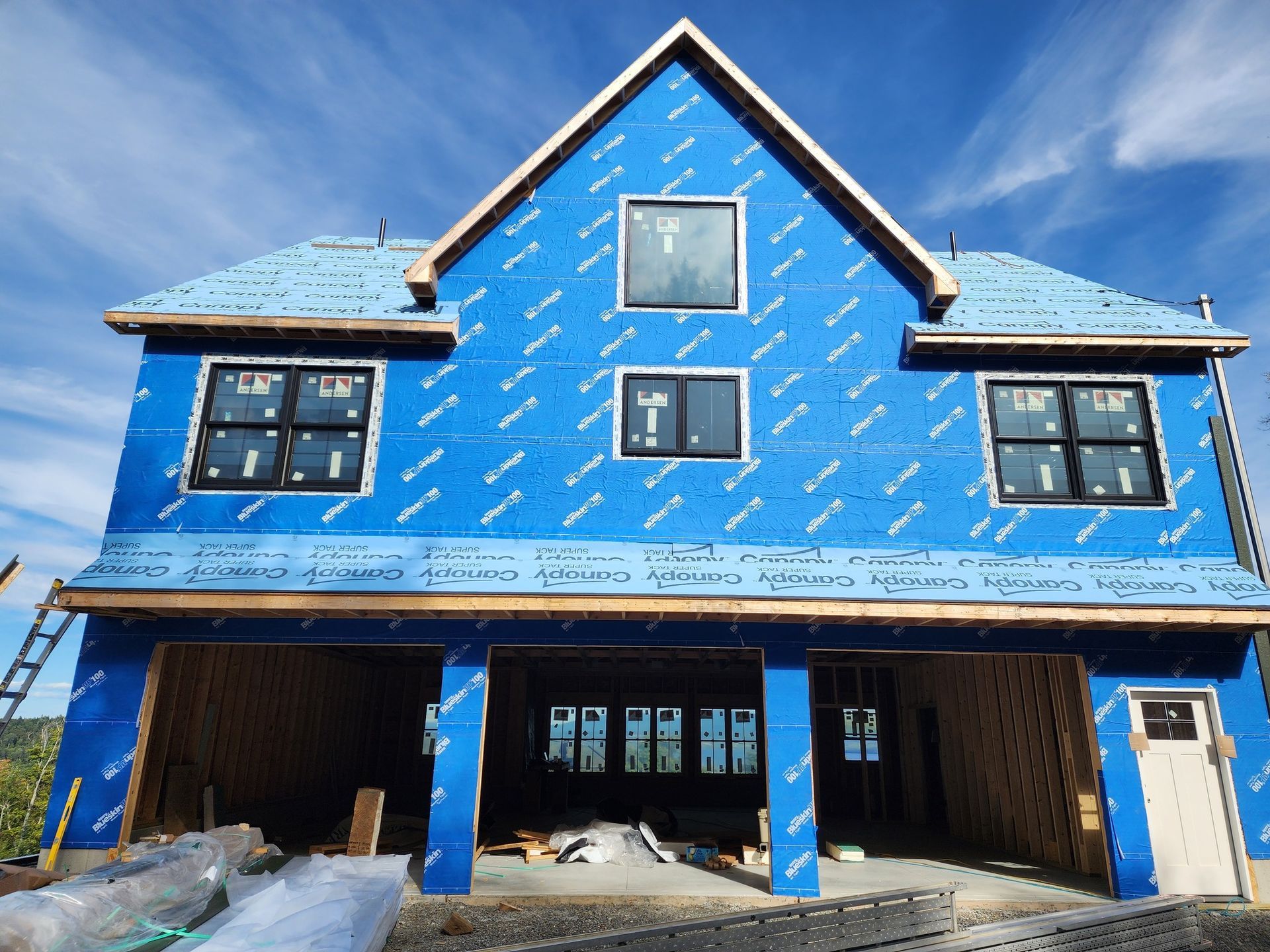 Two-story house under construction, blue protective wrap, black windows, garage, blue sky.