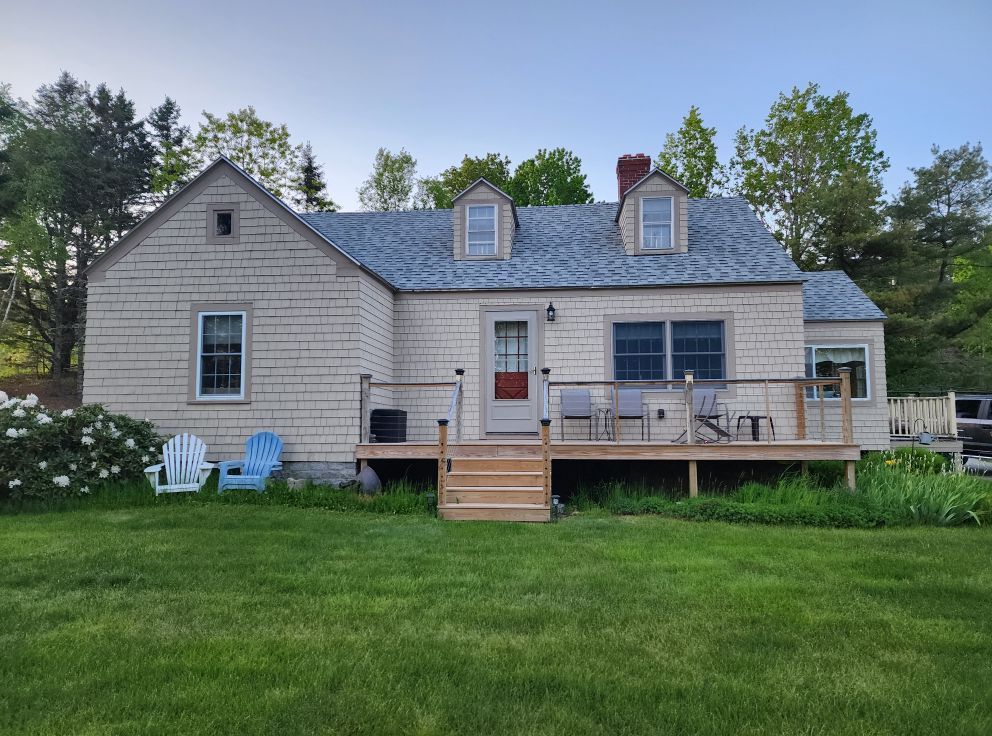 Cottage with beige siding, gray roof, wooden deck, two dormers, and green lawn. Three chairs on the lawn.