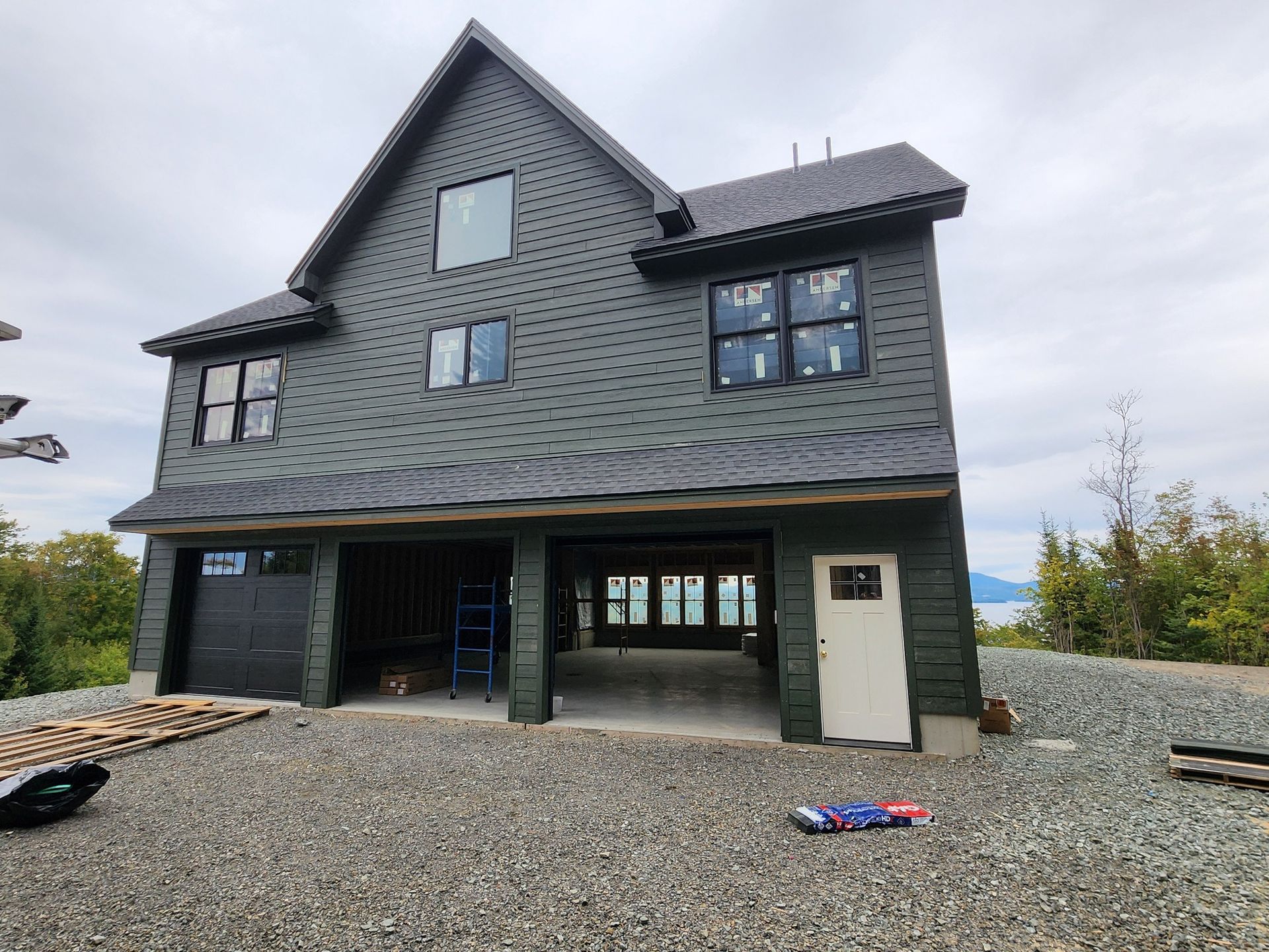 Two-story dark green house under construction with an open garage, on a gravel lot.