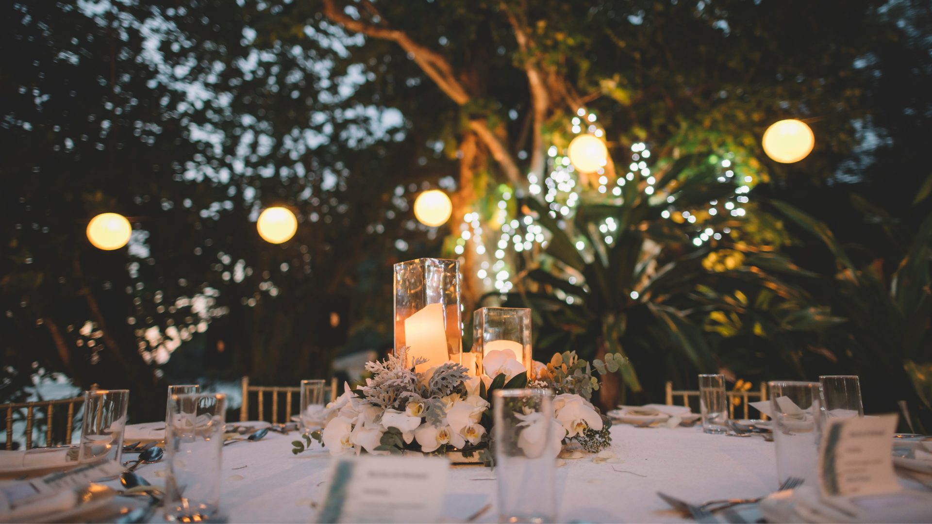 Elegant outdoor dinner table with candles, white flowers, and warm string lights in a garden setting
