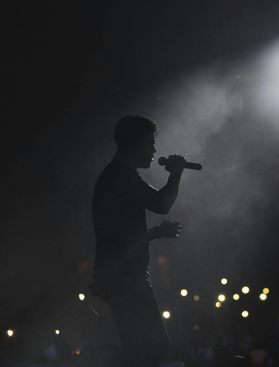 Two musicians performing onstage under warm orange lights, one singing into a microphone while playing guitar.