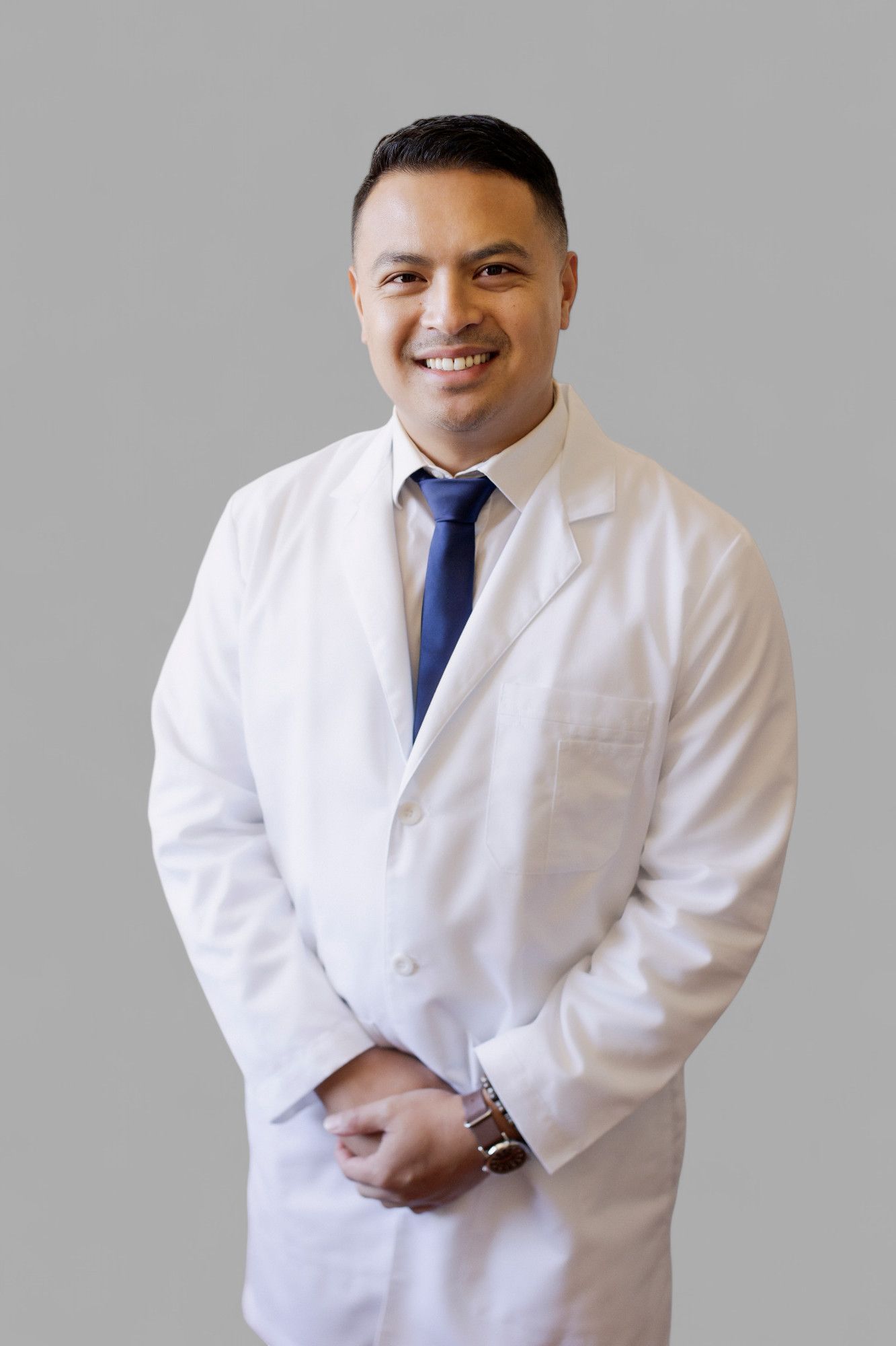 A smiling professional in a white lab coat and blue tie, posed against a solid gray background.