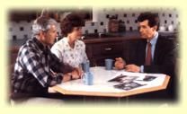 Three people sit around a table in a meeting, reviewing papers and talking in a bright office.