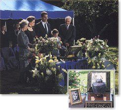 Outdoor funeral service with floral arrangements and people standing beside a casket draped in blue