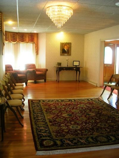 Spacious living room with chandelier, patterned rug, chairs, and sunlight through curtained windows