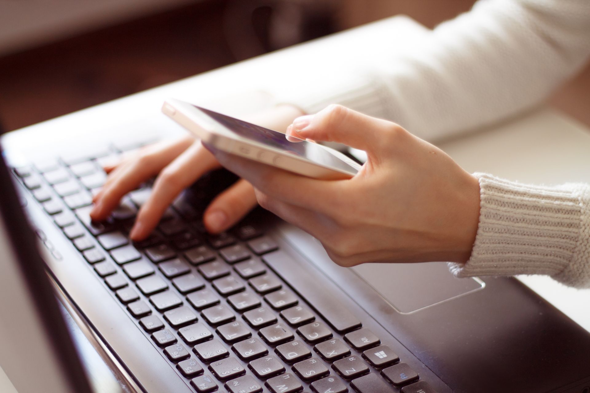 Hands typing on a laptop while holding a smartphone above the keyboard.