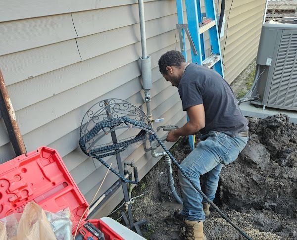 A person in jeans works on a gas line outside a building, near tools and an AC unit.