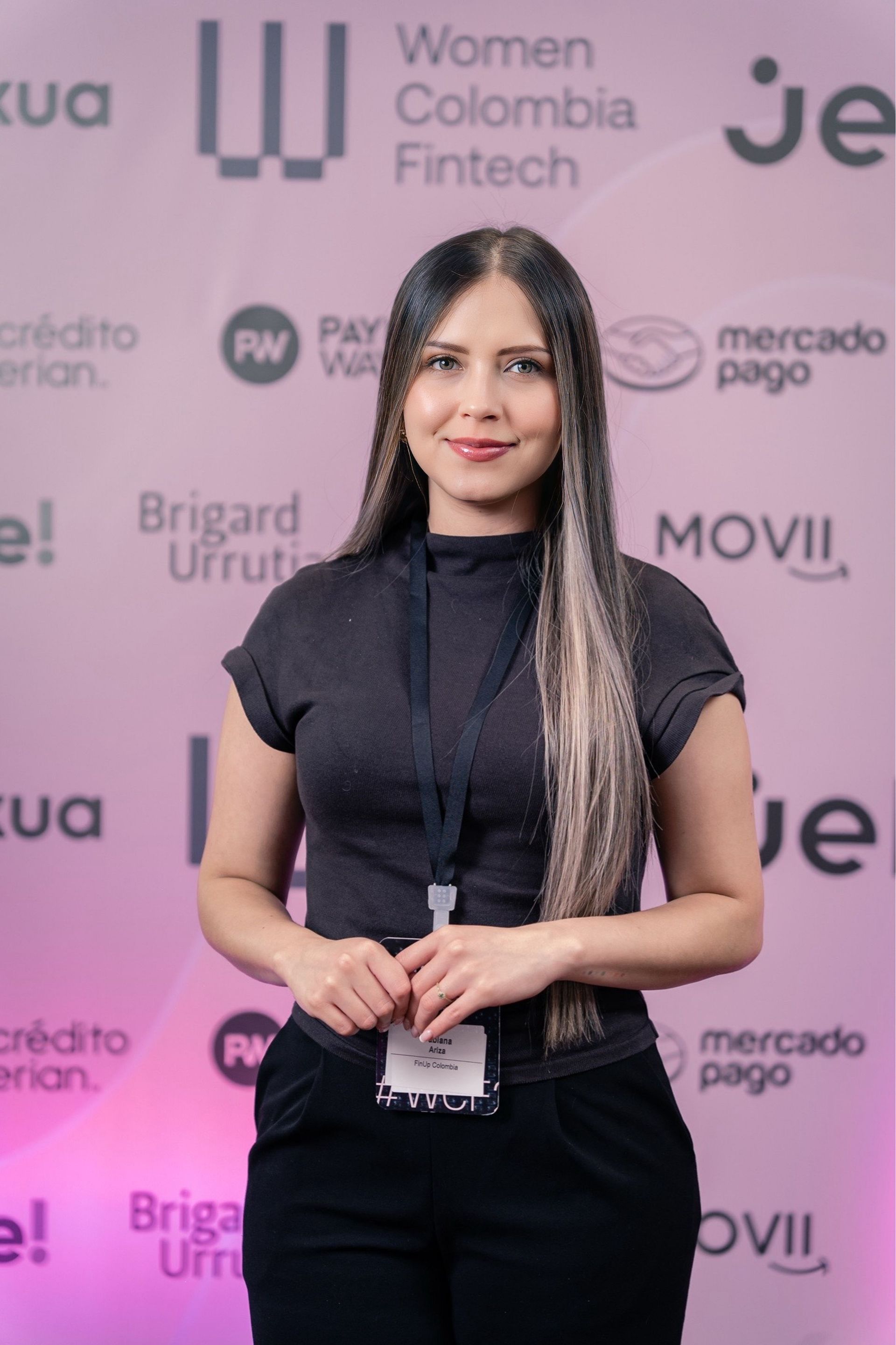Mujer de pie en un evento de Women Colombia Fintech, vestida de negro, frente a un fondo rosa de un patrocinador.