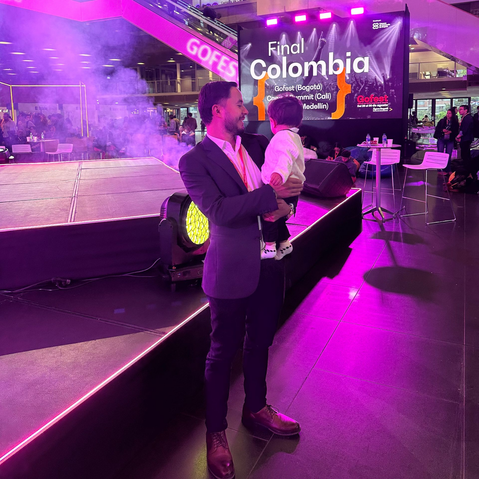 Un hombre habla en un escenario iluminado con luces moradas en un evento con un letrero que dice