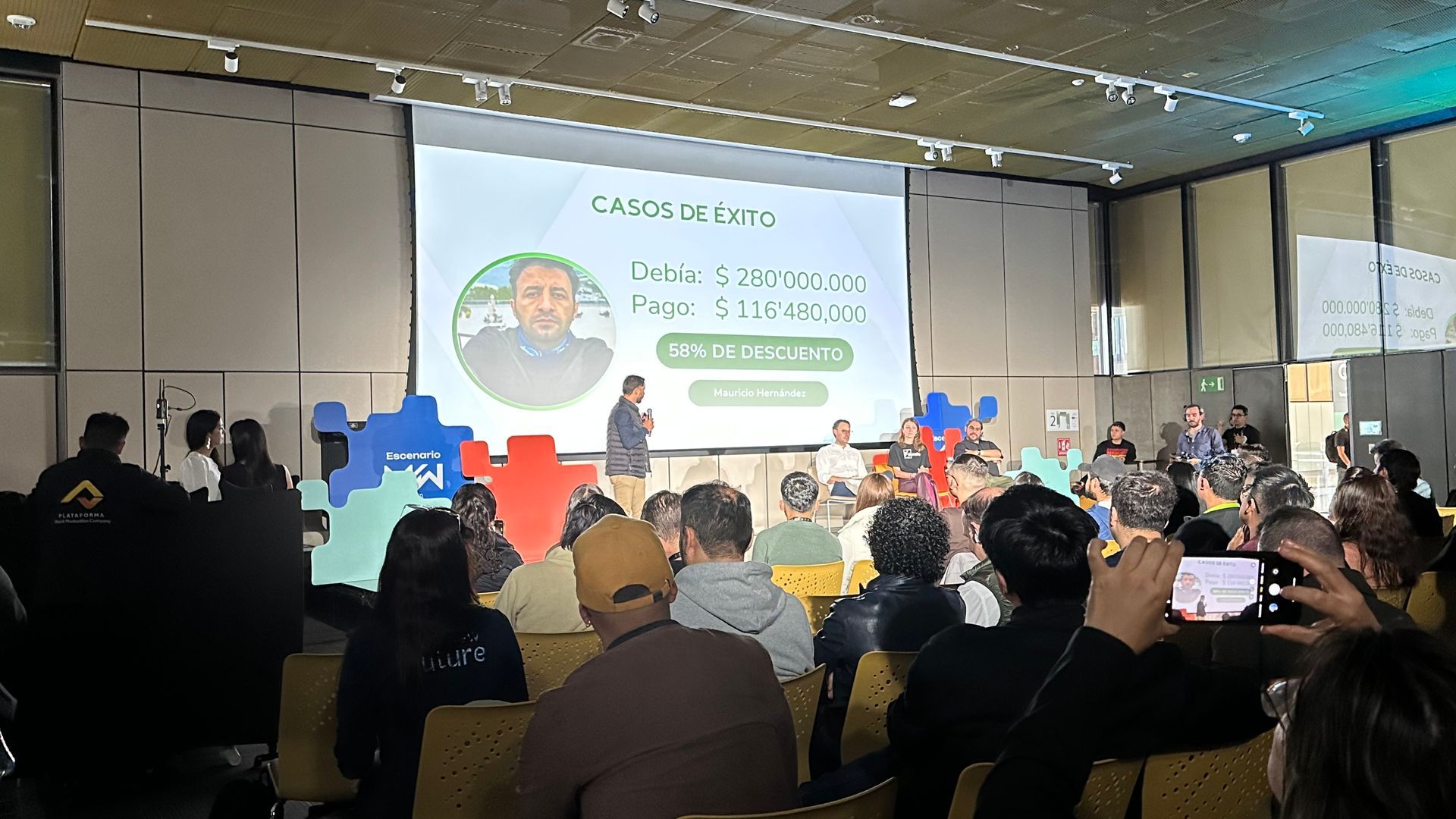 El público observa una colorida presentación en el escenario de una sala de conferencias, con una diapositiva proyectada y oradores sentados.
