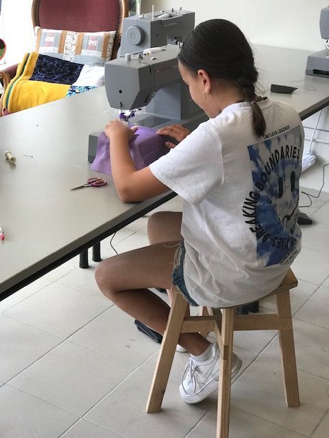 Een persoon in een wit T-shirt naait stof met een naaimachine aan een witte tafel.