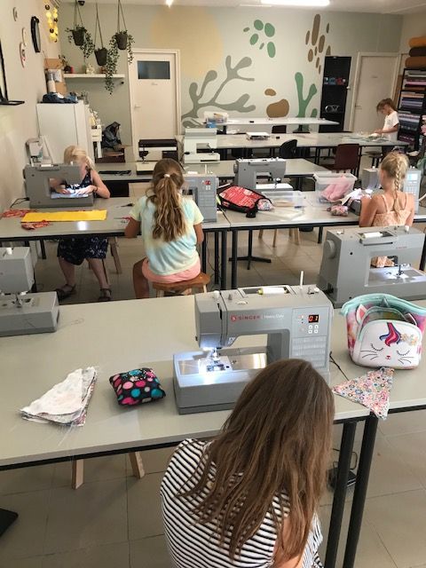 Kinderen naaien aan individuele tafels in een helder verlicht klaslokaal, met behulp van Singer-naaimachines.