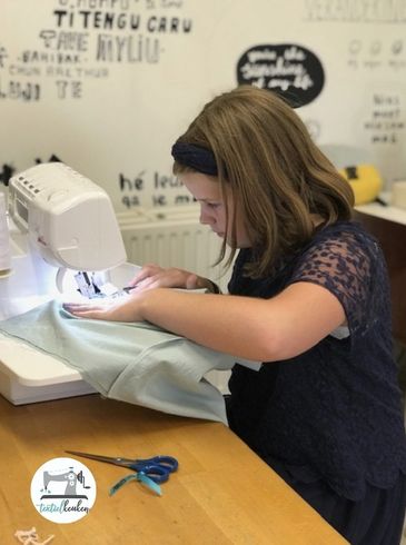 Meisje naait op een naaimachine aan een houten tafel. Schaar in de buurt. Lichtblauwe stof.